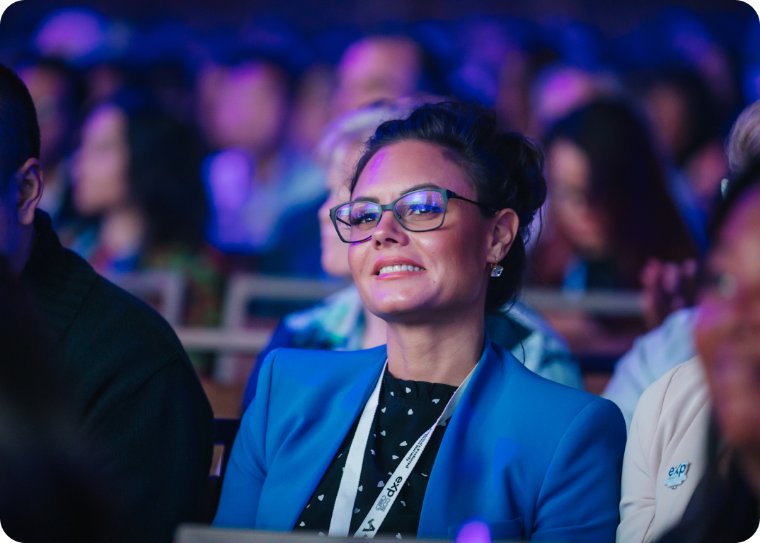 A woman in glasses and a blue blazer, seated and smiling at a conference event.