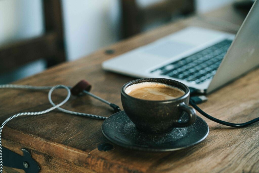 A close-up of a coffee cup on a saucer, placed on a wooden desk next to a laptop and a cable.