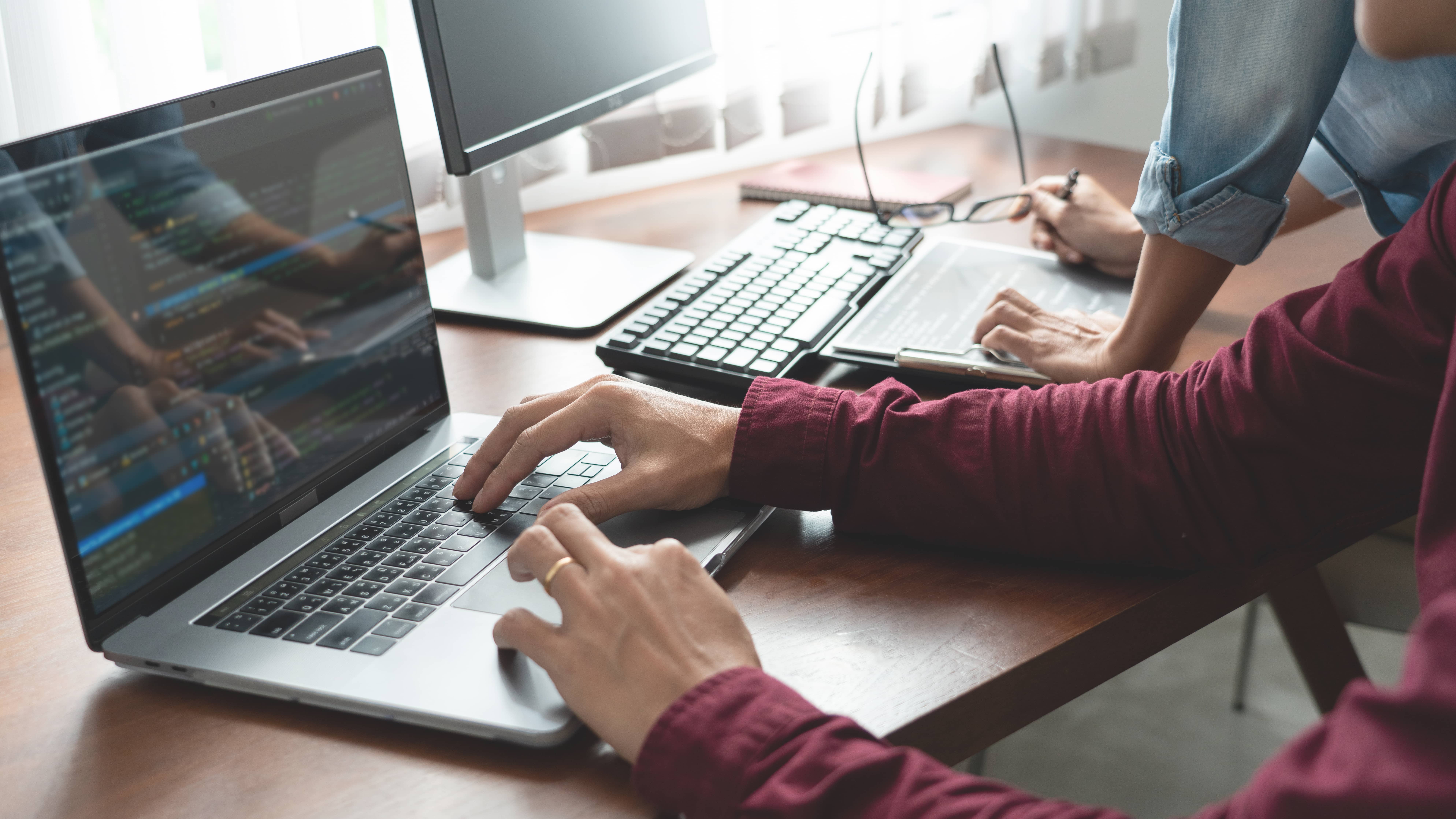 A person typing on a laptop with dual monitors in a work setting.