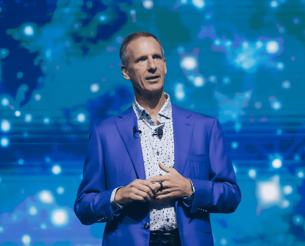 eXp world holdings CEO Glenn Sanford in a blue blazer and speaking on stage, holding his hands together, with a background of blue digital and abstract patterns.